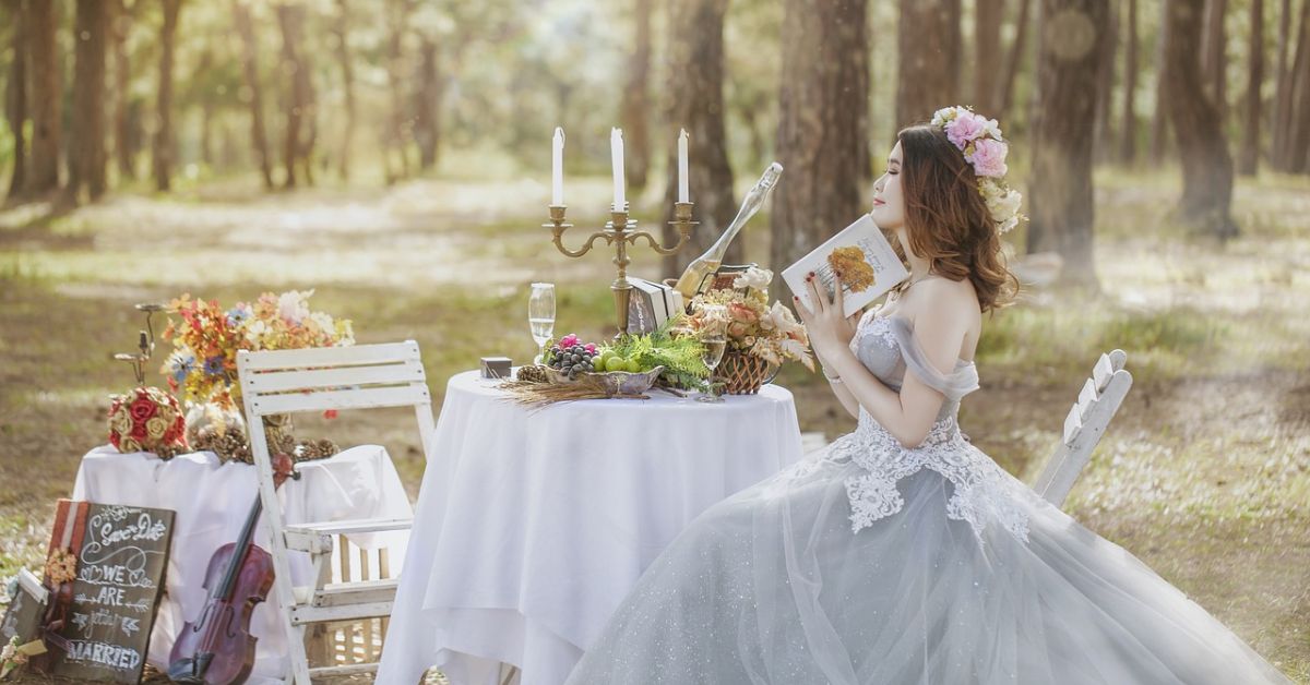 A woman in a wedding dress sits at a rustic table surrounded by trees, embodying the charm of outdoor weddings.