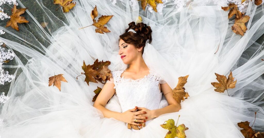 A woman in an Old Money wedding dress lies gracefully on the ground, surrounded by vibrant autumn leaves.