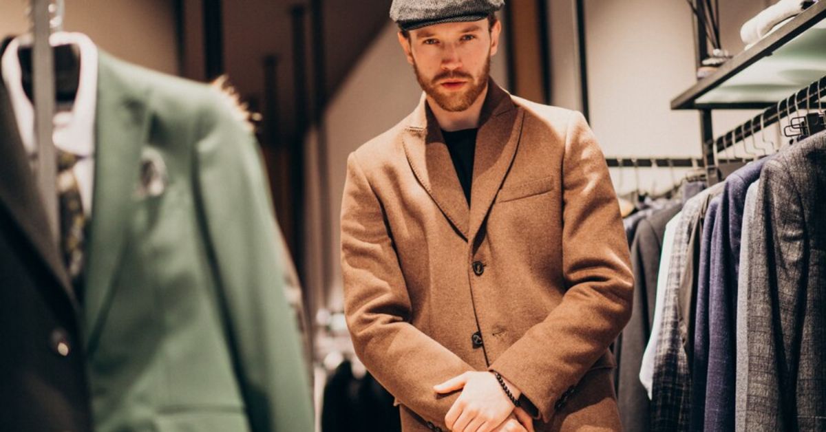 A man in a hat and coat stands confidently before a rack of classic, quality clothing, embodying old money aesthetics.