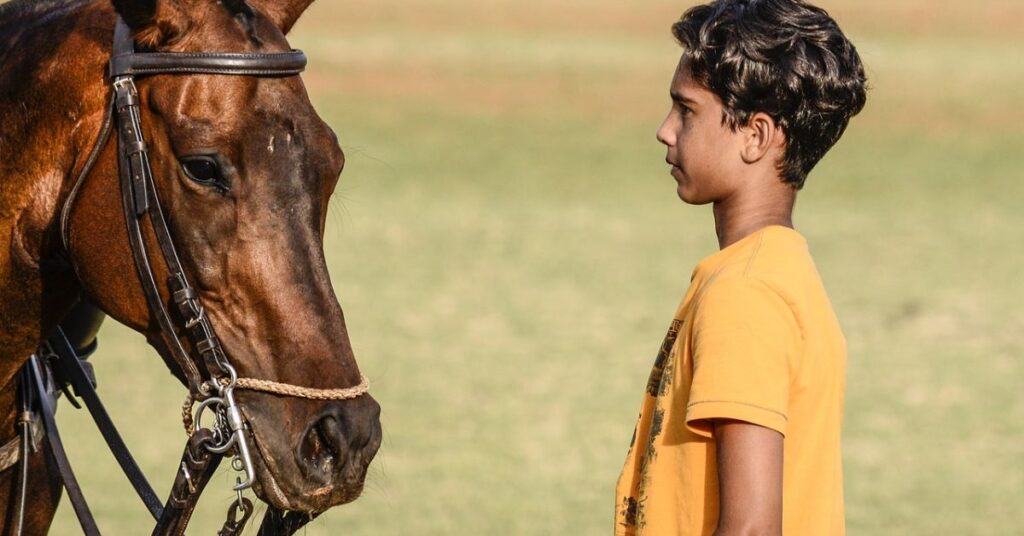 A young boy stands beside a horse, embodying the classic elegance of old money style in a polo setting.