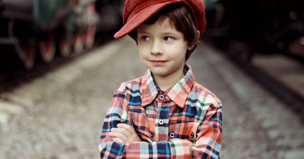 A young boy in a red hat smiles as he stands in front of a train, embodying a classic charm with his old money vibe.