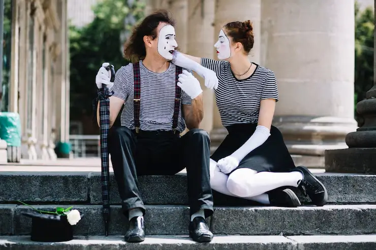 Two street performers dressed in mime costumes sit on stone steps, wearing matching black-and-white striped tops, black pants and skirts, white gloves, and face paint, styled in a playful yet coordinated 1950s rockabilly fashion.