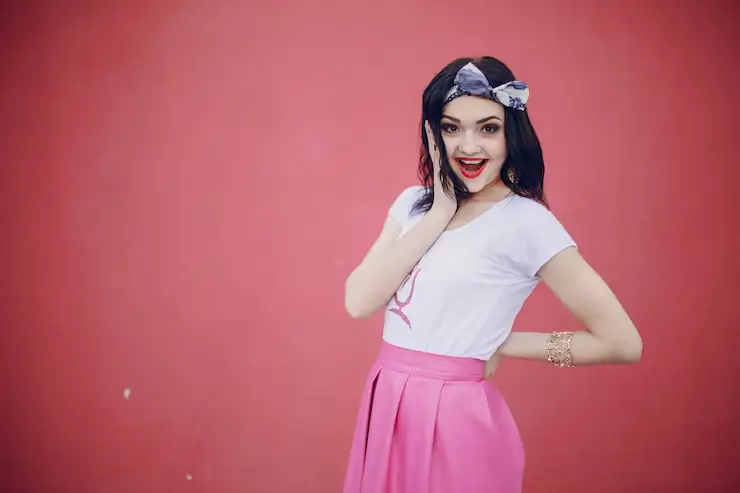 Smiling woman in 1950s rockabilly fashion wearing a white graphic T-shirt, high-waisted pink skirt, and a vintage-style headband, posing against a vibrant pink background.